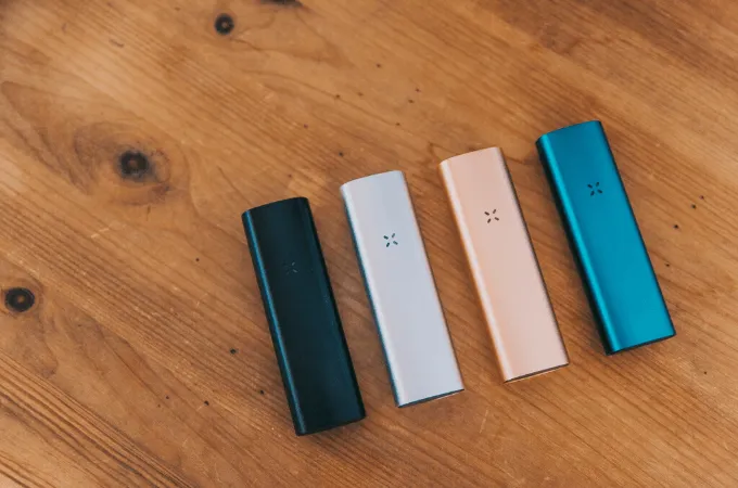 Four PAX vaporizers in teal, white, rose gold and blue on a wooden table